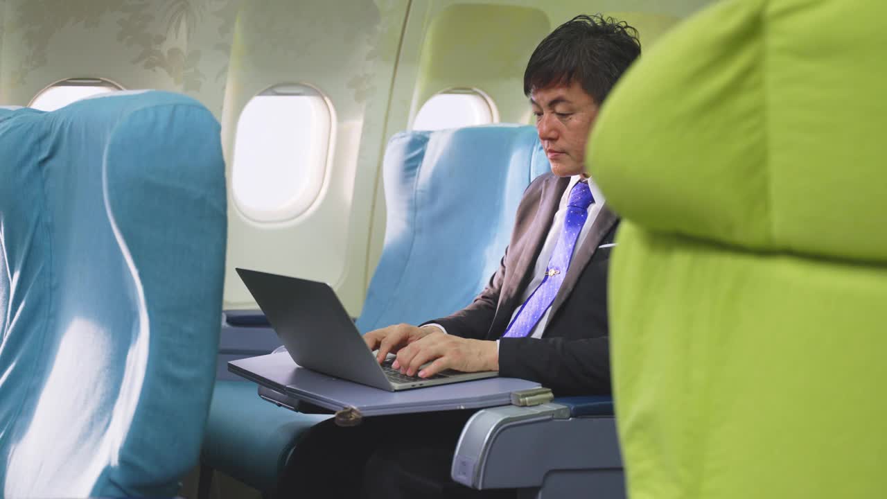 一位亚洲高管商人坐在商务舱，在飞机上用笔记本电脑工作。这个人很忙，眼睛感到很累。关注医疗保健和技术为商务人士的理念。