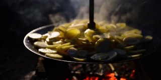 土豆在火上炸，有人在煎锅下煽火。土豆是在明火上的平底锅里煎的。晚上在户外做饭。为游客和旅行者提供野餐、露营。