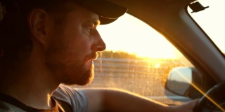男人在日落时开车。男性手放在方向盘上，靠近。一个大胡子男人在高速公路上开车，目不转睛地盯着路面。从驾驶室里面看侧面。阳光照在挡风玻璃上。