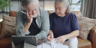 退休的亚洲老夫妇在沙发上一起检查和计算财务账单，参与财务文书工作，在客厅用电子银行笔记本电脑在线纳税
