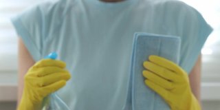 戴黄色手套的女人在洗窗户。她把喷雾瓶里的水洒在玻璃上，用蓝色抹布擦干净。关注前景中的水滴。家务和清洁公司员工的工作概念