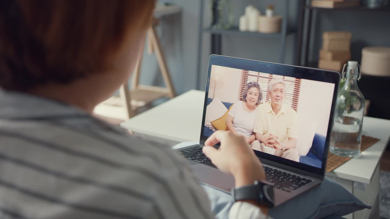 年轻的亚洲女孩使用笔记本电脑视频电话会议与年长的家庭爸爸和妈妈，而工作在家坐在客厅的沙发上。