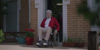 蓄着胡须的残疾人在门廊上用慢动作推着轮椅欣赏户外的阳光。宽幅照片的快乐男性白人残疾退休在夏天春天的一天。