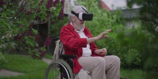 戴着VR头盔的残疾老人坐在户外轮椅上，用慢动作微笑着移动双手。积极快乐的残疾白人退休享受增强现实在后院的春天花园。