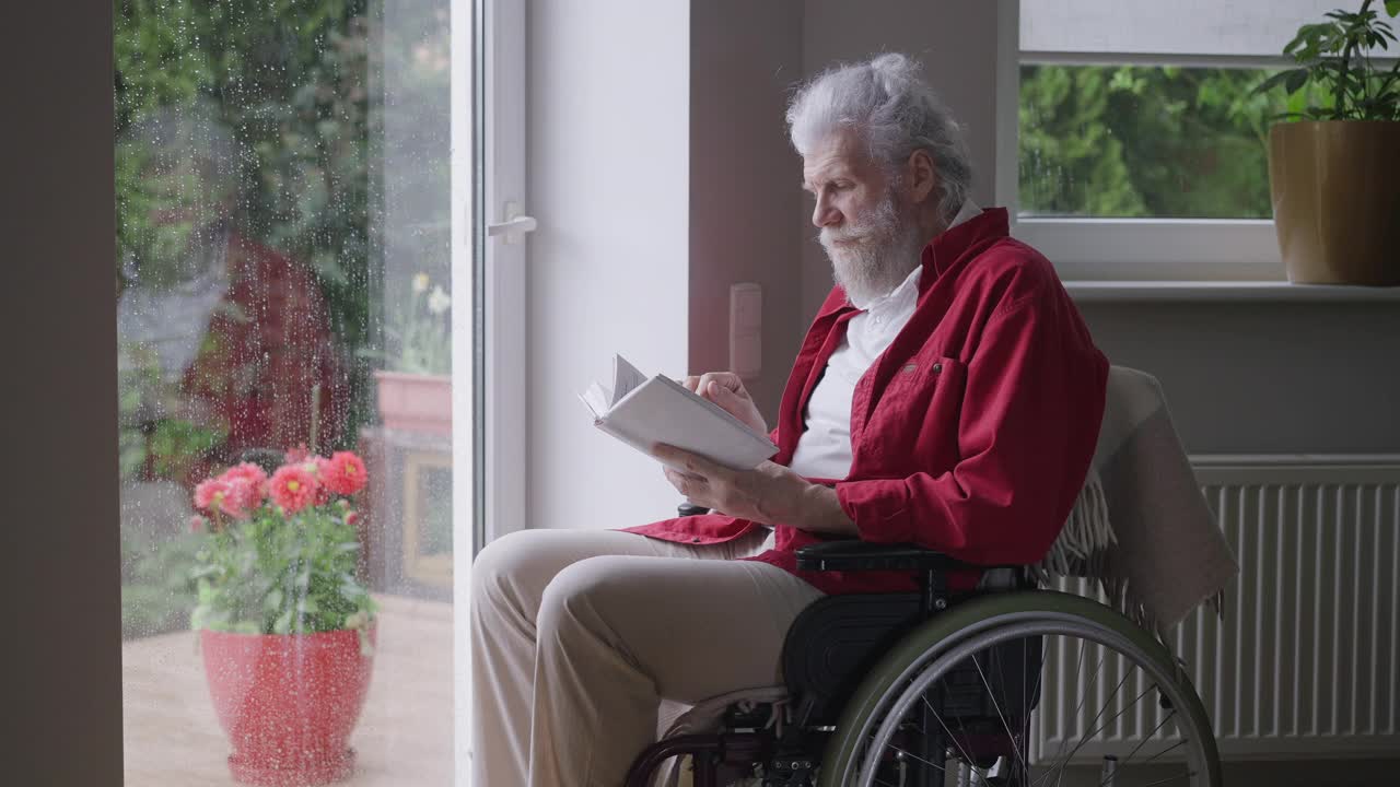 一名坐在轮椅上的老年残疾人在下雨天看着窗外看书。智能残疾的白人退休老人在家里享受文学的肖像。缓慢的运动。