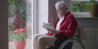 一名坐在轮椅上的老年残疾人在下雨天看着窗外看书。智能残疾的白人退休老人在家里享受文学的肖像。缓慢的运动。