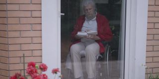 自信聪明英俊的老头坐在轮椅上看书合着书想着坐在家里的玻璃门后面。在室内用宽镜头拍摄的聪明体贴的白人退休老人。缓慢的运动。