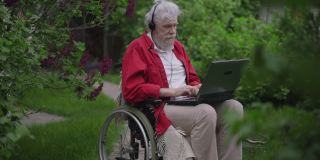 英俊开朗的残疾老人坐在轮椅上微笑着在笔记本电脑上视频聊天。留着大胡子的白人退休老人在后院的花园里聊天。现代技术。