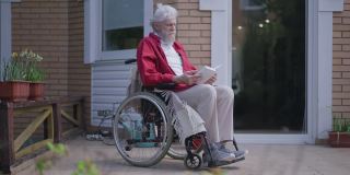 有胡子的残疾人老人边看边合上书，看着阳光。户外后院门廊上，坐在轮椅上的聪明的白人残疾退休老人的广角照片。