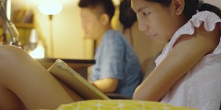 亚洲女孩晚上在家里用电脑玩网络游戏的哥哥旁边用平板电脑看电影，生活理念。