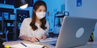 快乐亚洲女商人戴着医用口罩，在新常态下保持社交距离，以预防病毒，在办公室晚上使用笔记本电脑回到工作岗位。