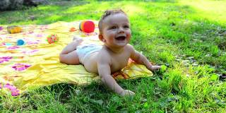 刚出生的光着身子蹒跚学步的婴儿，小男孩躺在铺着毯子的草地上，旁边放着玩具，笑着，向上看