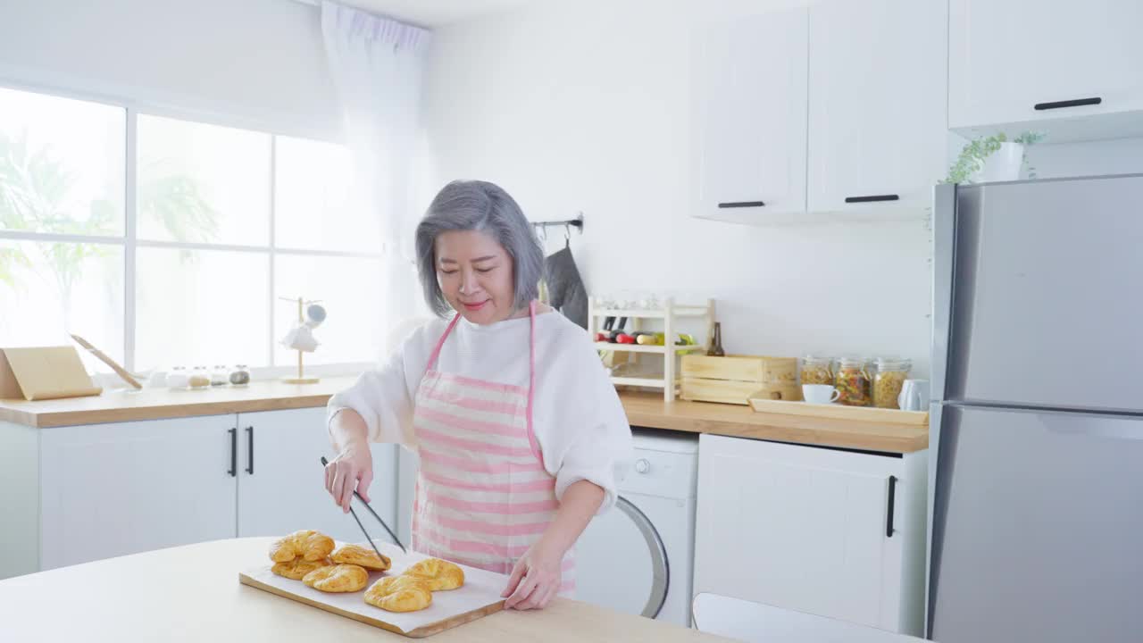 亚洲可爱的家庭，年轻的女儿看着老妈妈在厨房做饭。美丽的女性享受打发闲暇时光，抱着年迈的妈妈在家里的餐桌上烤牛角面包。活动的关系