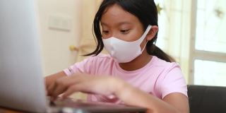 亚洲女孩戴着医用口罩在家里用电脑笔记本电脑观看在线视频内容，居家隔离和新常态生活理念