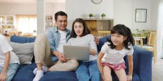 亚洲家庭爸爸和妈妈坐在沙发上享受笔记本电脑上的网上购物，而女儿和儿子在家里客厅的沙发上边玩边大喊大叫。