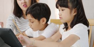 快乐亚洲家庭家庭教育，母亲教孩子在家里的客厅使用数码平板电脑。