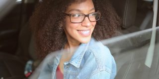 快乐的年轻微笑的非洲裔美国女人黑头发戴眼镜的司机坐在新的棕色汽车，微笑着看着相机享受旅程。驾驶课程和人寿保险概念
