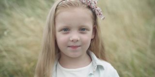 特写照片的自信美丽的小白种人金发女孩与灰色的眼睛看着相机微笑。快乐放松可爱的孩子在户外与春秋草的背景。