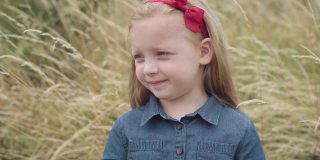 可爱迷人的小女孩，金色头发上系着红丝带，站在秋天的田野里望着远方。漂亮的高加索孩子的肖像享受闲暇。童年和自然的概念。