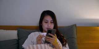 孤独的年轻亚洲女人使用智能手机和社交媒体盖着毯子在卧室