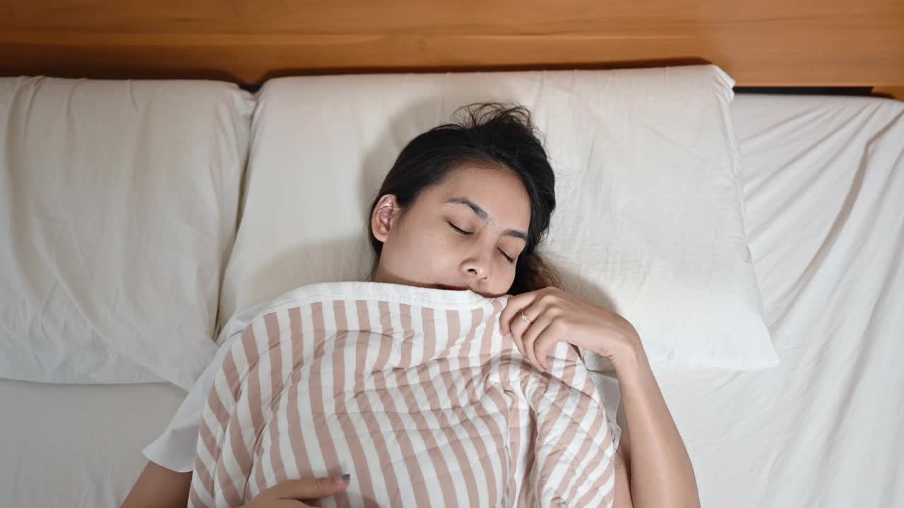 年轻的亚洲女人醒来，伸展手臂在床上的毯子在早上