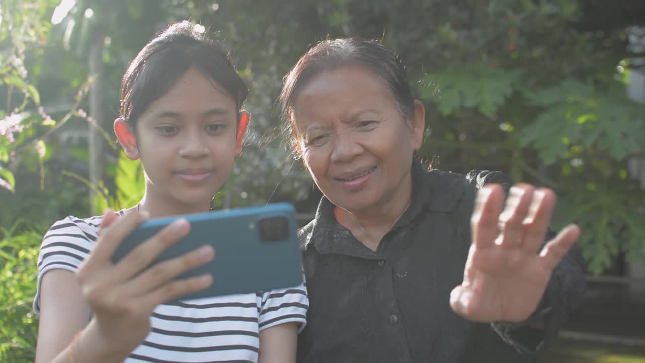 亚洲可爱的十几岁女孩和她的奶奶喜欢在家里的后院用手机视频聊天，一边挥手一边和家人聊天。