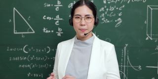 年轻的亚裔女教师戴着耳机与学生进行视频会议。女教师在课堂上培训数学黑板在线课程。
