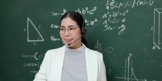 年轻的亚洲女教师手持粉笔，戴着耳机，与学生看着摄像机进行视频会议。女教师在课堂上培训数学黑板在线课程。