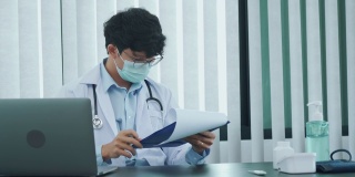 一位亚洲医生正在用笔记本电脑工作，在办公室里写病人的病史。