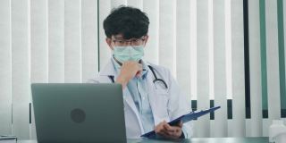 一位亚洲医生正在用笔记本电脑工作，在办公室里写病人的病史。
