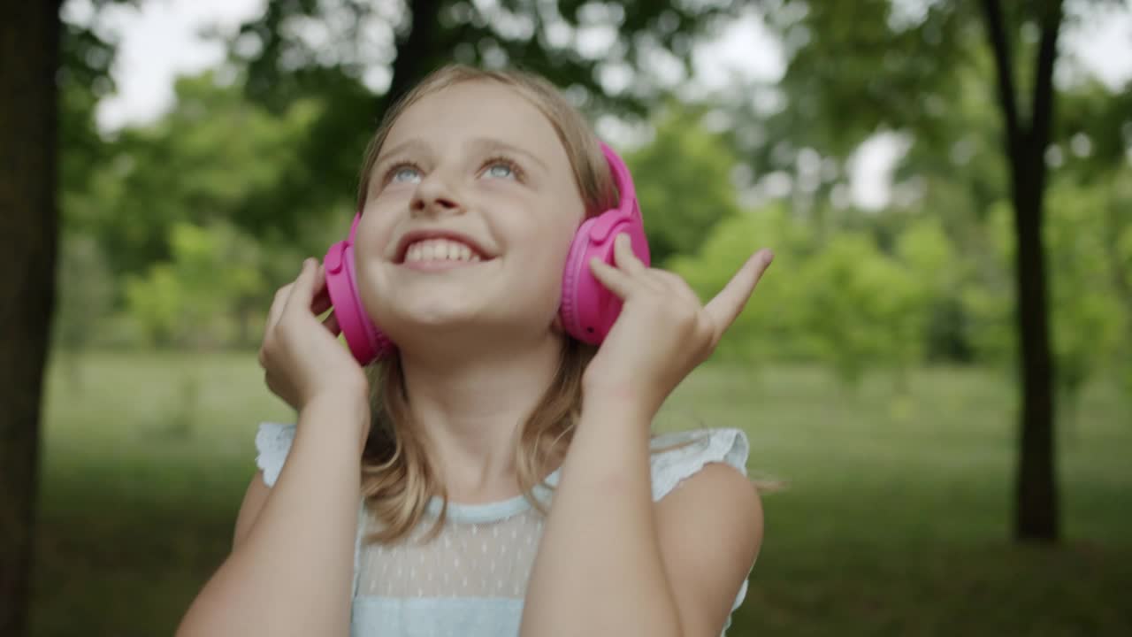 快乐的少年在公园里戴着时髦的粉色耳机听着音乐，抬起头来