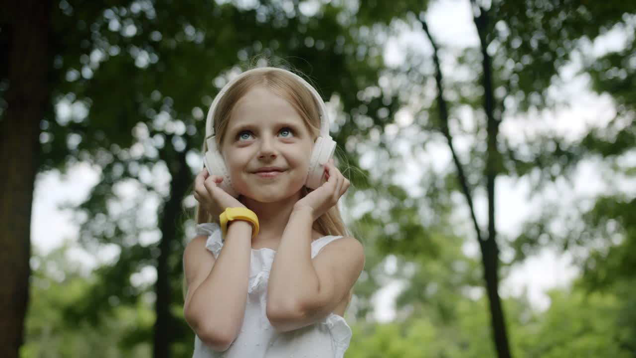 快乐的孩子在公园里用现代的白色耳机听音乐，向上看