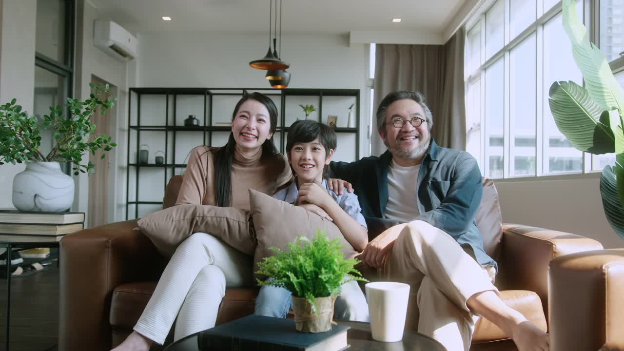 幸福甜蜜的亚洲家庭爸爸妈妈和儿子坐在沙发上看电影从电视在家里。笑笑亚洲家庭居家隔离隔离时刻在封锁国家秩序。