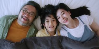 幸福亚洲可爱的一家人一起躺在床上，笑亚洲父母和儿子在毯子下看着摄像机俯视图家的家庭概念