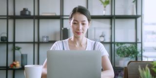 亚洲女性的妻子穿着休闲布在家工作，带着笔记本电脑，专注于餐桌的家庭背景，亚洲女性在家工作
