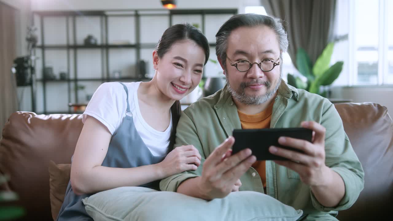 离开的亚洲夫妇在家里客厅的沙发沙发上一起玩智能手机在线移动游戏，周末的休闲居家隔离生活方式居家室内背景