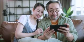 离开的亚洲夫妇在家里客厅的沙发沙发上一起玩智能手机在线移动游戏，周末的休闲居家隔离生活方式居家室内背景