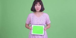 工作室拍摄的美丽怀孕的亚洲妇女拿着数字平板电脑，在绿色的工作室背景下微笑和思考