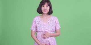 摄影棚拍摄了一名美丽的亚洲孕妇，在绿色的摄影棚背景下微笑并竖起大拇指