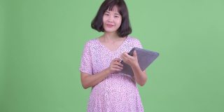 工作室拍摄美丽的怀孕亚洲妇女微笑和使用数字平板电脑在绿色的工作室背景