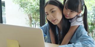 一位亚洲母亲带着她可爱的小女儿在家工作。当妈妈在笔记本电脑上工作时，一个亚洲小孩从后面拥抱她的妈妈。