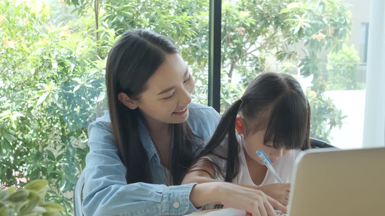 一位亚洲母亲喜欢用笔记本电脑教她可爱的女儿做作业。亚洲小女孩喜欢和她漂亮的妈妈一起学习。