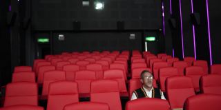 一位留着白胡子的亚裔中国男子坐在空荡荡的电影院的红色座位上一边吃一边看电影。