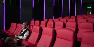 一位留着白胡子的亚裔中国男子坐在空荡荡的电影院的红色座位上一边吃一边看电影。