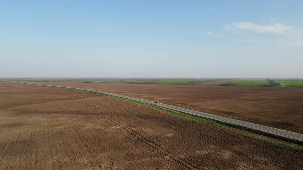 灰色的长沥青公路，汽车在棕色的田野之间，在蓝色的春日天空下，云朵鸟瞰
