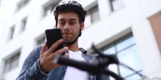特写脸严肃的年轻人戴着防护头盔，用手机坐在自行车上，从下面拍摄。