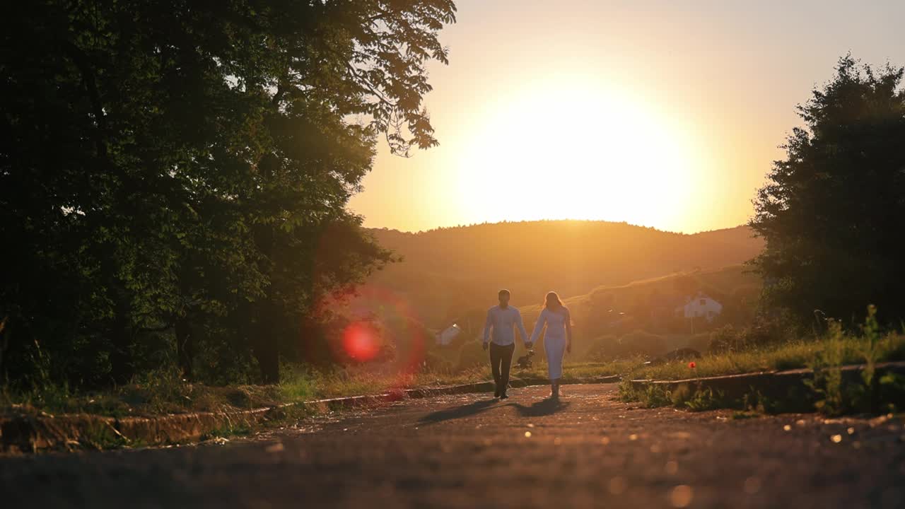 年轻夫妇手牵着手在夕阳下散步。一对相爱的情侣在夕阳下的公园散步。橙色的日落