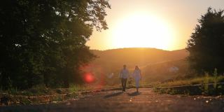 年轻夫妇手牵着手在夕阳下散步。一对相爱的情侣在夕阳下的公园散步。橙色的日落