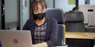 亚洲女雇员戴着口罩使用笔记本电脑在商业办公室工作与同事保持社交距离保护冠状病毒或covid-19爆发大流行。职场新常态政策
