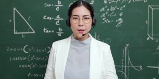 年轻的亚裔女教师戴着耳机与学生进行视频会议。女教师在课堂上培训数学黑板在线课程。